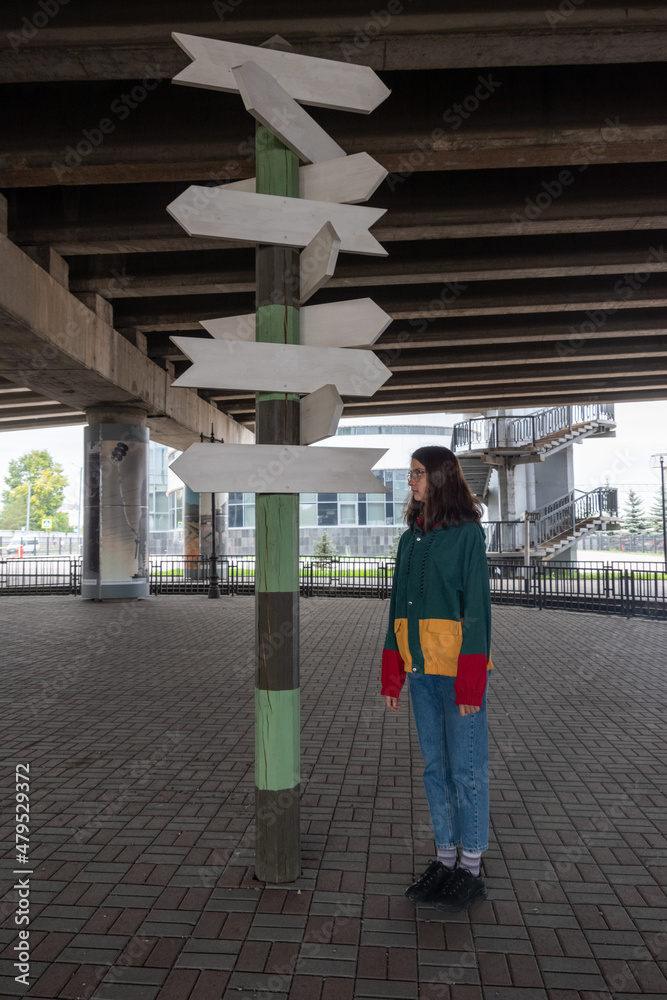 Obraz premium A teenage girl in multicolored trendy clothes stands confusedly near a sign with empty arrows in opposite directions