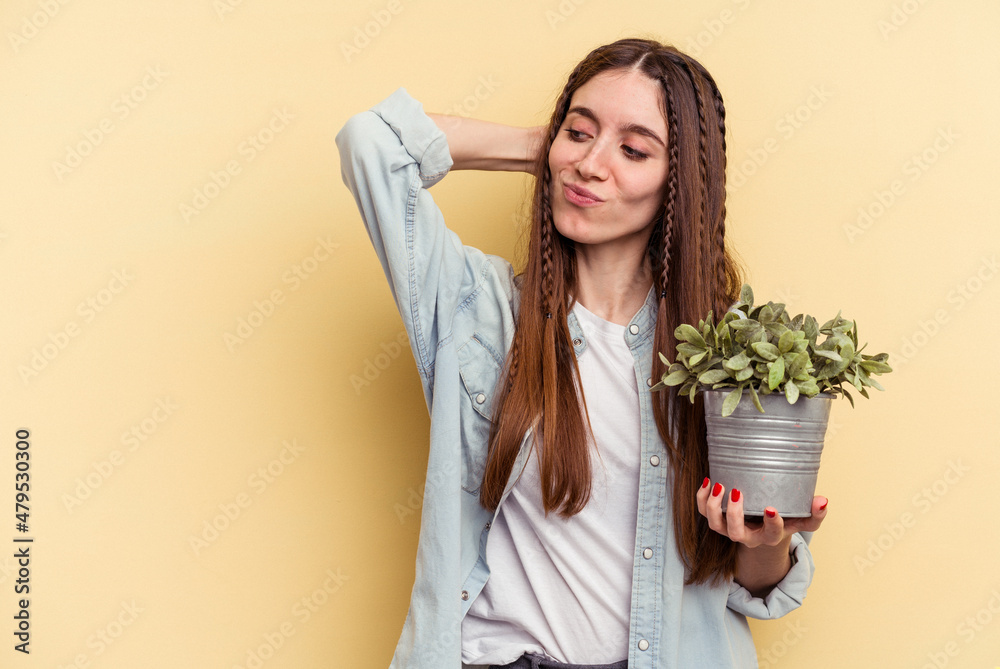 Young caucasian woman holding a plant isolated on yellow background touching back of head, thinking and making a choice. Young caucasian woman holding a plant isolated on yellow background touching back of head, thinking and making a choice.