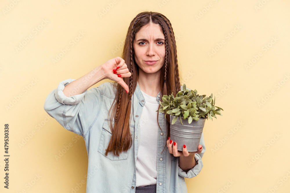 Young caucasian woman holding a plant isolated on yellow background showing a dislike gesture, thumbs down. Disagreement concept. Young caucasian woman holding a plant isolated on yellow background showing a dislike gesture, thumbs down. Disagreement concept.