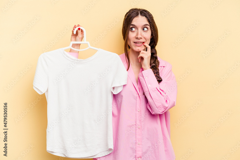 Young caucasian woman holding a hanger isolated on yellow background relaxed thinking about something looking at a copy space. Young caucasian woman holding a hanger isolated on yellow background relaxed thinking about something looking at a copy space.