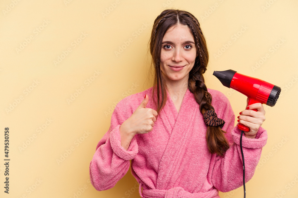 Young woman holding a hairdryer isolated on yellow background smiling and raising thumb up Young woman holding a hairdryer isolated on yellow background smiling and raising thumb up