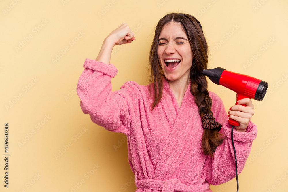 Young woman holding a hairdryer isolated on yellow background raising fist after a victory, winner concept. Young woman holding a hairdryer isolated on yellow background raising fist after a victory, winner concept.
