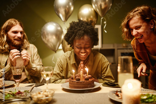 African happy woman sitting at the table with birthday cake with candles and laughing  she celebrating birthday with friends at home