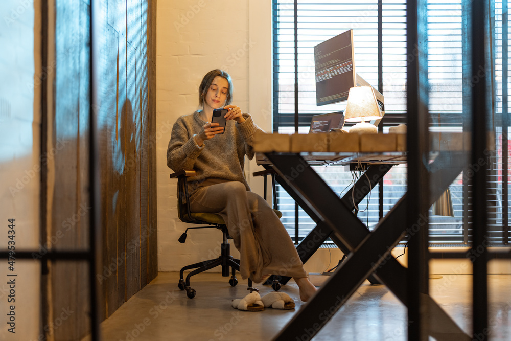 Young woman using phone while sitting at workplace with laptop and desktop computers at cozy home office interior. Concept of freelance and remote work at home. Woman wearing domestic clothes Young woman using phone while sitting at workplace with laptop and desktop computers at cozy home office interior. Concept of freelance and remote work at home. Woman wearing domestic clothes