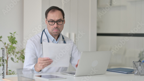 Middle Aged Doctor with Laptop reading Documents in Clinic