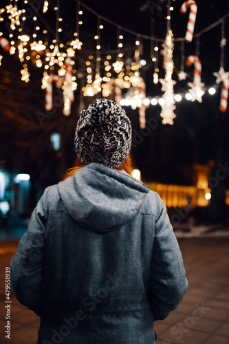 Outdoor night photo of young beautiful happy smiling girl enjoying festive decoration, posing in Christmas fair, in street of european city, wearing knitted beanie hat