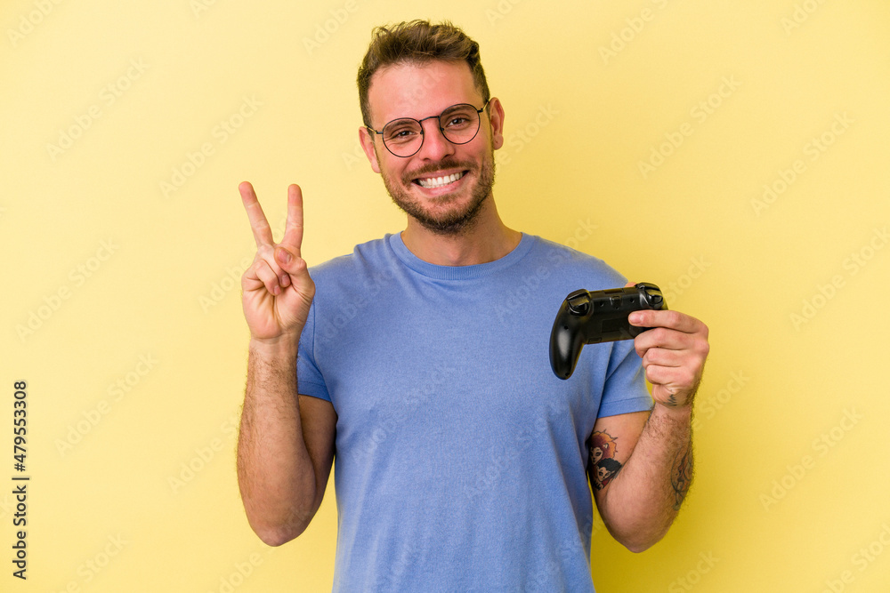 Young caucasian man holding a game controller isolated on yellow background joyful and carefree showing a peace symbol with fingers. Young caucasian man holding a game controller isolated on yellow background joyful and carefree showing a peace symbol with fingers.