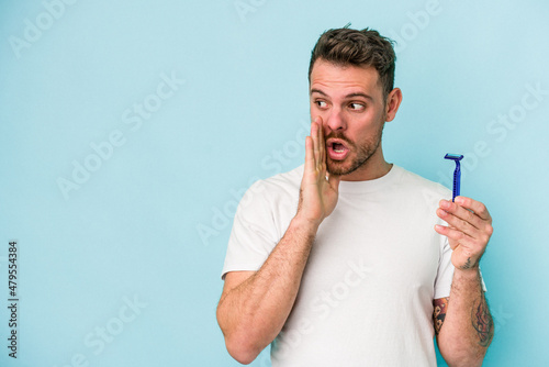 Young caucasian man shaving his beard isolated on blue background is saying a secret hot braking news and looking aside