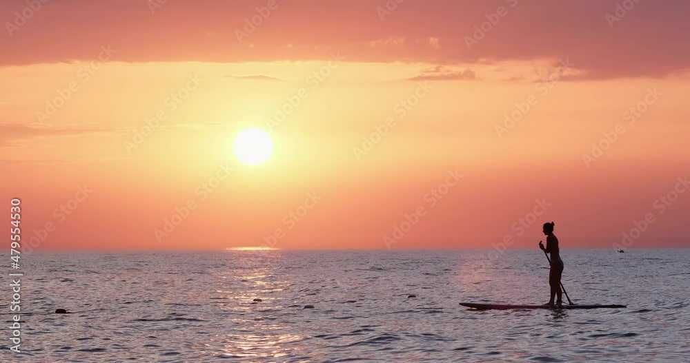 Athletic young woman wearing bikini swimsuit swims standing on SUP enjoying ripple sea view at bright colorful sunset on cloudy sky Athletic young woman wearing bikini swimsuit swims standing on SUP enjoying ripple sea view at bright colorful sunset on cloudy sky