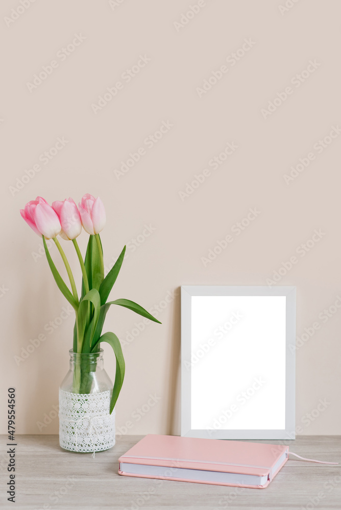Home interior with decorative elements. Layout with a white frame and pink tulips in a vase and a pink notebook on the table on a light beige background