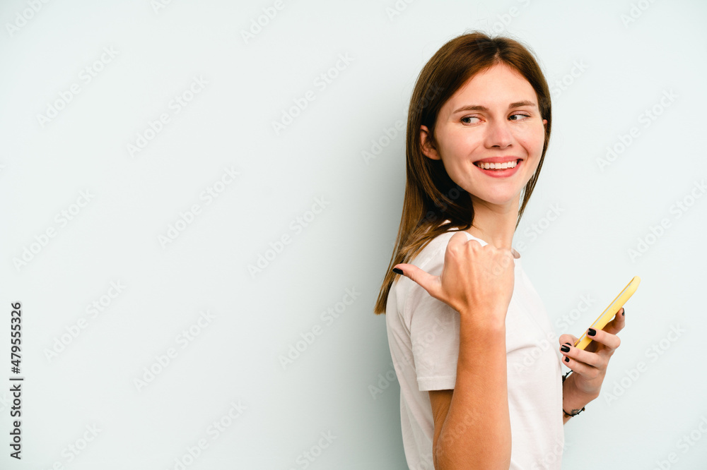 Young English woman holding mobile phone isolated on blue background points with thumb finger away, laughing and carefree. Young English woman holding mobile phone isolated on blue background points with thumb finger away, laughing and carefree.
