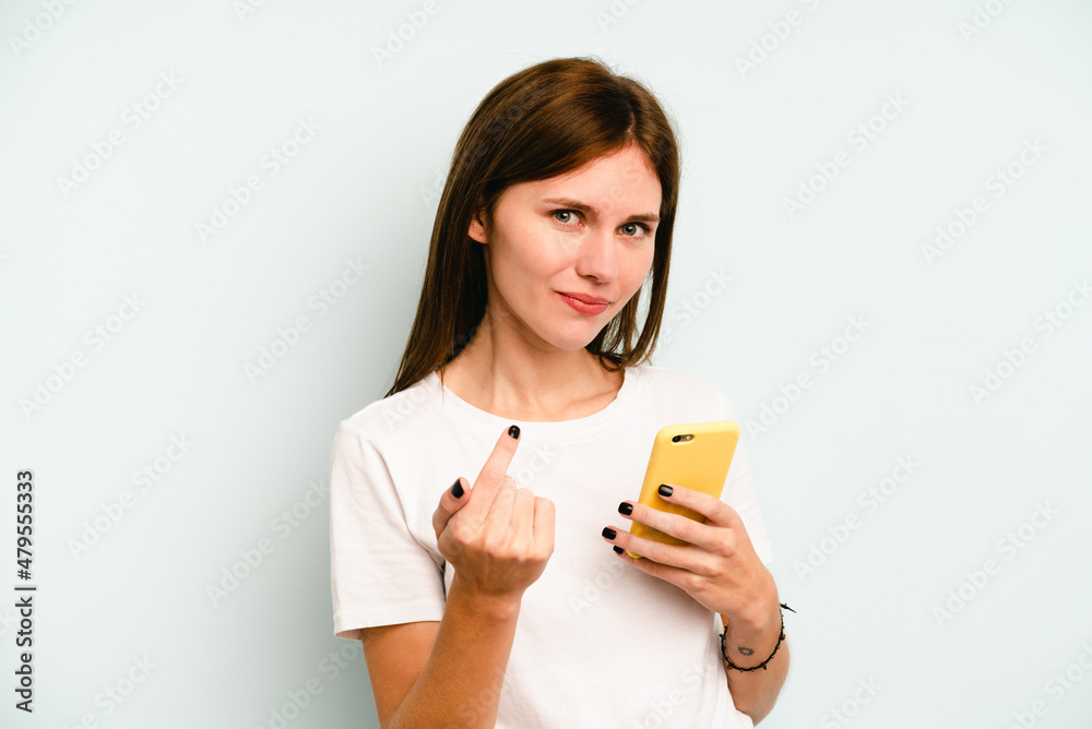 Young English woman holding mobile phone isolated on blue background pointing with finger at you as if inviting come closer. Young English woman holding mobile phone isolated on blue background pointing with finger at you as if inviting come closer.