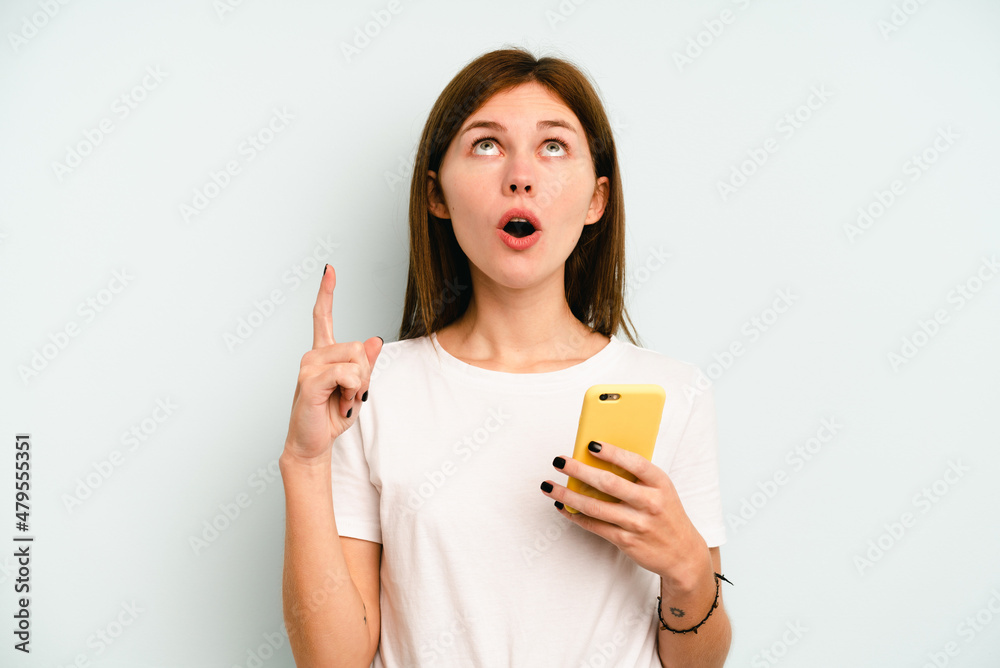 Young English woman holding mobile phone isolated on blue background pointing upside with opened mouth. Young English woman holding mobile phone isolated on blue background pointing upside with opened mouth.