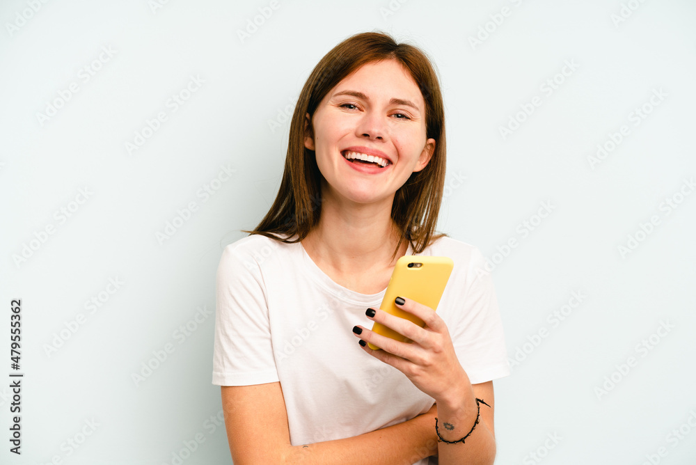 Young English woman holding mobile phone isolated on blue background laughing and having fun. Young English woman holding mobile phone isolated on blue background laughing and having fun.