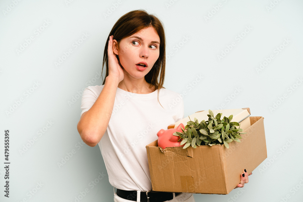 Young English woman making a move while picking up a box full of things isolated on blue background trying to listening a gossip. Young English woman making a move while picking up a box full of things isolated on blue background trying to listening a gossip.