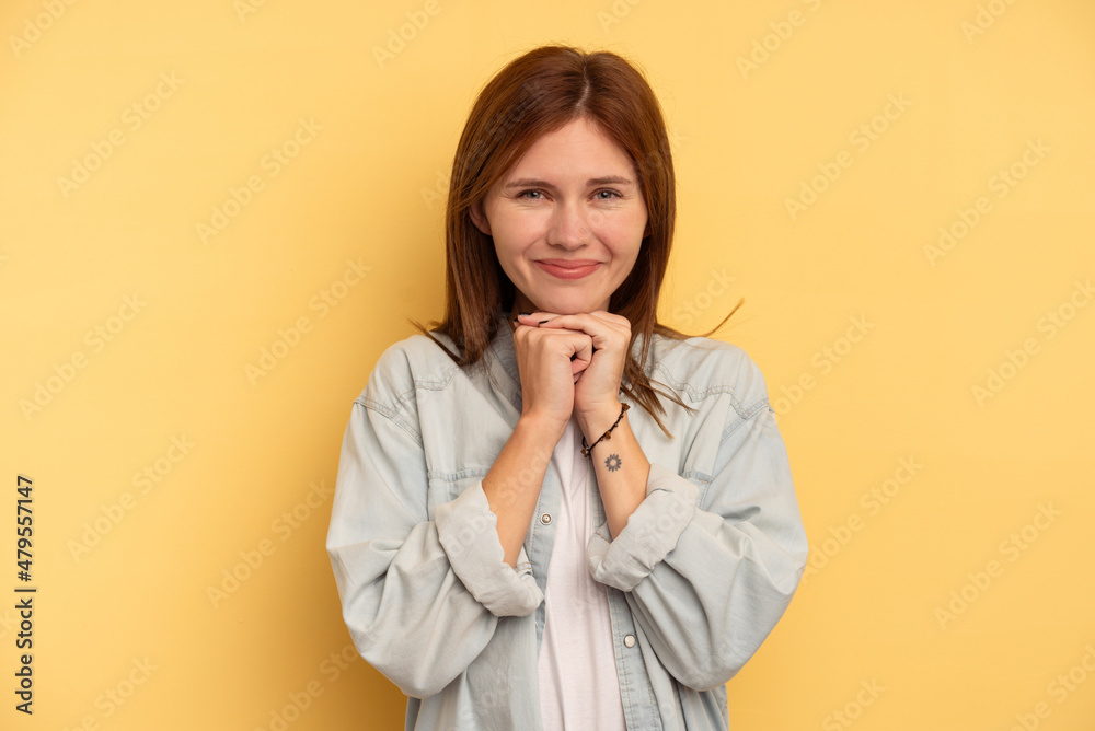 Young English woman isolated on yellow background keeps hands under chin, is looking happily aside. Young English woman isolated on yellow background keeps hands under chin, is looking happily aside.