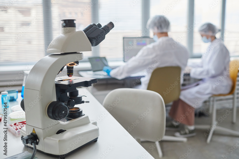 Close-up of microscope on the table in the laboratory with two scientists in the background Close-up of microscope on the table in the laboratory with two scientists in the background