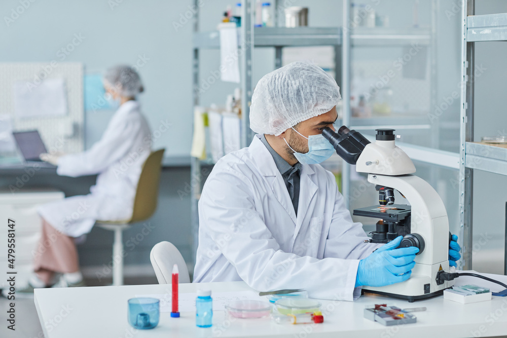 Young scientist in lab coat and protective mask examining sample of new virus with microscope during work in the laboratory Young scientist in lab coat and protective mask examining sample of new virus with microscope during work in the laboratory