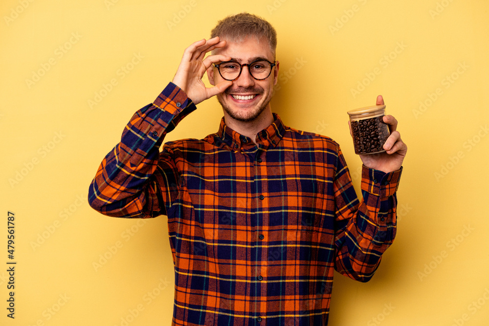 Young caucasian man holding coffee jar isolated on yellow background excited keeping ok gesture on eye. Young caucasian man holding coffee jar isolated on yellow background excited keeping ok gesture on eye.