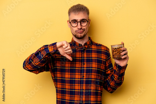 Young caucasian man holding a almond jar isolated on yellow background showing a dislike gesture  thumbs down. Disagreement concept.