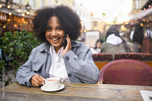 Fashion street style portrait of attractive young natural beauty African American woman with afro hair in tweed jacket speaks phone outdoors in sidewalk cafe. Happy lady with coffee cup in big city