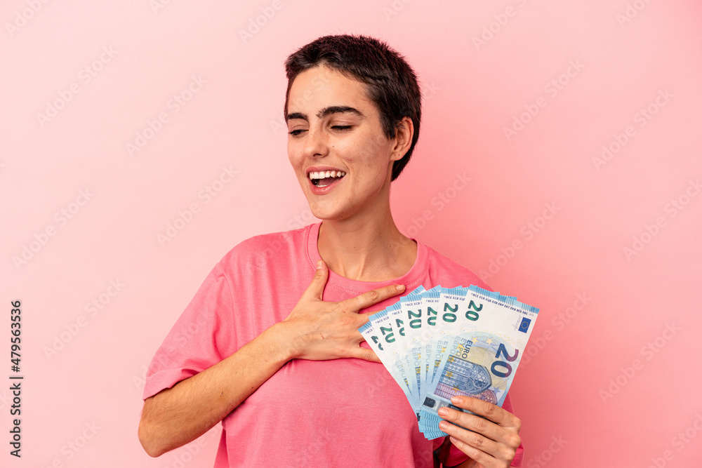 Young caucasian woman holding banknotes isolated on pink background laughs out loudly keeping hand on chest. Young caucasian woman holding banknotes isolated on pink background laughs out loudly keeping hand on chest.
