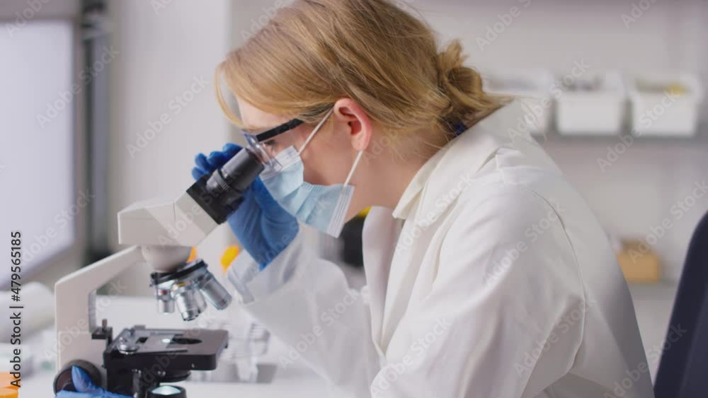 Female Lab Research Worker In PPE Looking Through Microscope And Recording Results On Laptop Female Lab Research Worker In PPE Looking Through Microscope And Recording Results On Laptop