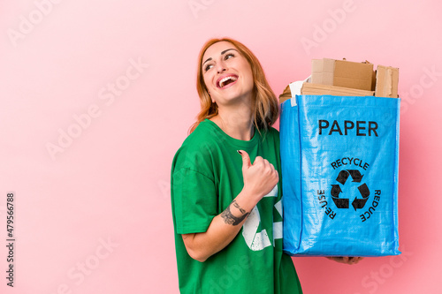 Young caucasian woman recycled paper isolated on pink background points with thumb finger away, laughing and carefree.