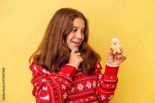 Little caucasian girl holding a Christmas cookies isolated on yellow background looking sideways with doubtful and skeptical expression.