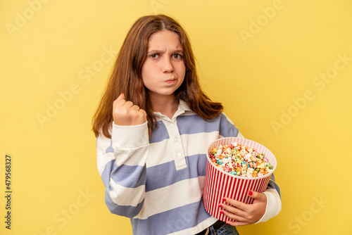 Little caucasian girl holding popcorns isolated on yellow background showing fist to camera, aggressive facial expression.