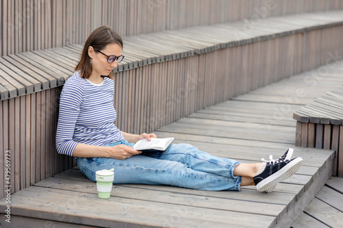 a young and confident European woman is resting and reading a book. a woman drinks coffee, relaxes or waits for a meeting with friends or colleagues in a park or a public place. meditation and reading