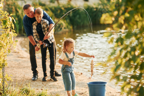 Girl putting fish into the bucket. Father with son and daughter on fishing together outdoors at summertime #479570583