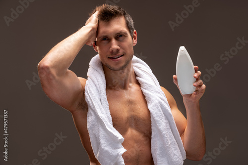 Portrait of young man smiling at camera, holding shampoo while preparing to applying product on his head isolated over brown background. Beauty, skincare, treatment concept