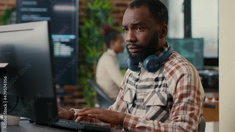 Portrait of focused african american programer wearing wireless headphones working looking at computer screen while typing. Software developer writing code in it agency office.