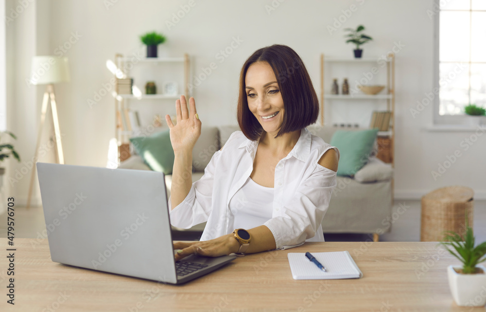 Friendly senior business woman waving in front of laptop webcam welcoming online conference participants. Woman enjoys virtual business communication and online negotiations sitting in her home office Friendly senior business woman waving in front of laptop webcam welcoming online conference participants. Woman enjoys virtual business communication and online negotiations sitting in her home office