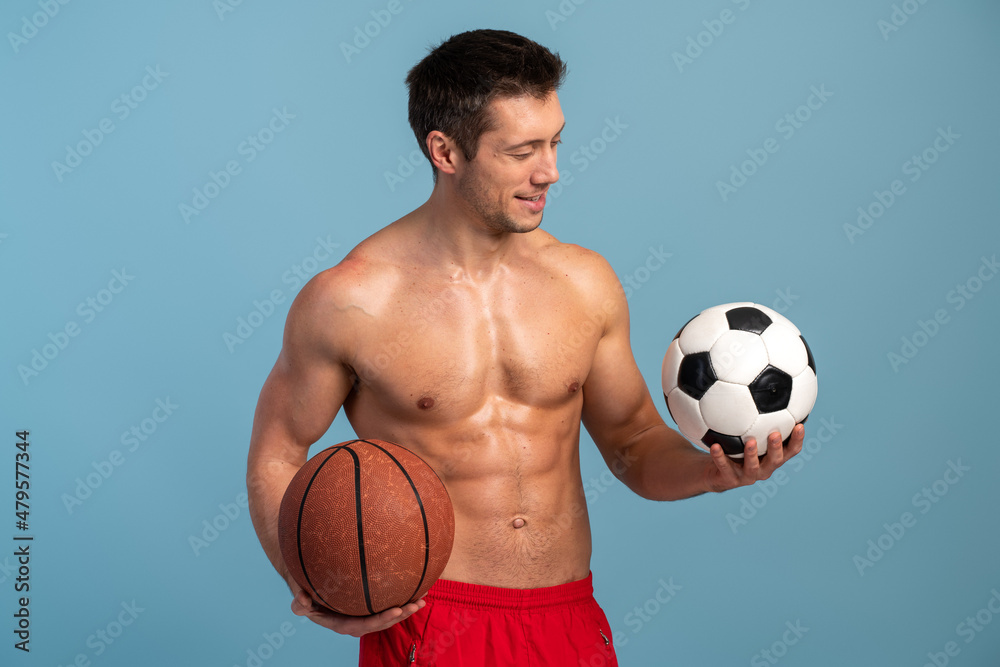 Waist up portrait view of the well shaped young man holding two balls and looking at the football ball over the light blue background Waist up portrait view of the well shaped young man holding two balls and looking at the football ball over the light blue background