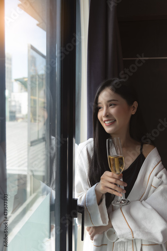 Happy woman in bathrobe with glass of champagne stand near window with happiness Drinking alcohol Asian sexy girl look through window for seeing nice view at hotel room with holding champagne glass