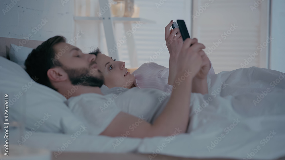Smiling woman using smartphone near blurred husband on bed.