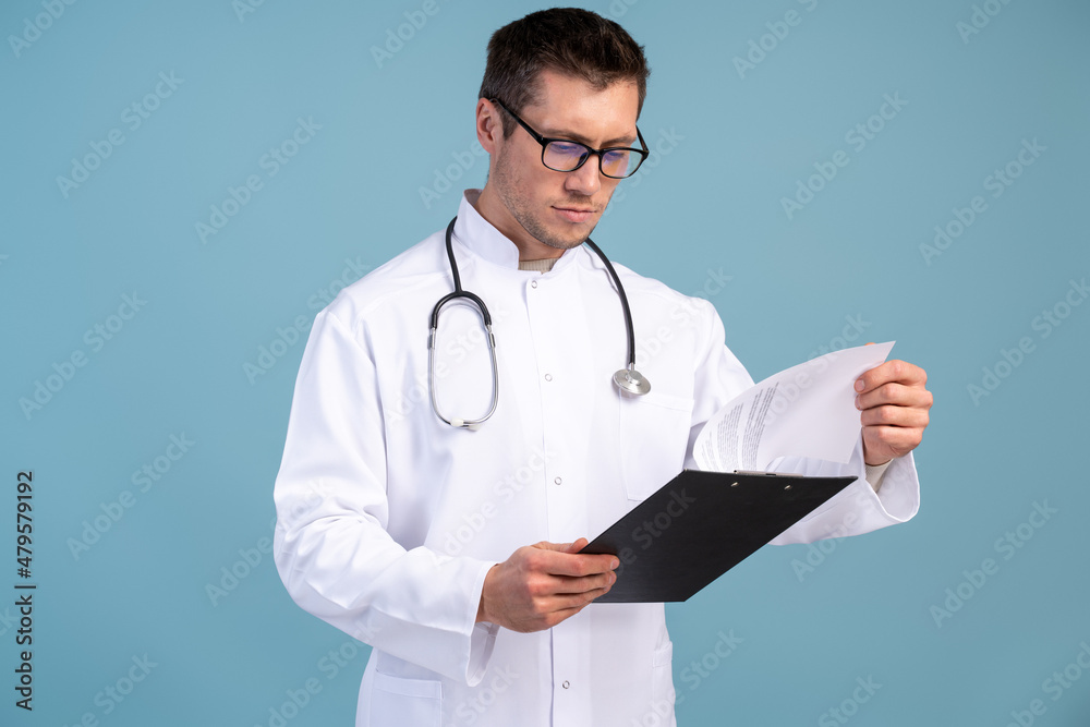 Waist up portrait view of the confident doctor in a uniform and in glasses holding folder and looking at it with serious face. Stock photo Waist up portrait view of the confident doctor in a uniform and in glasses holding folder and looking at it with serious face. Stock photo