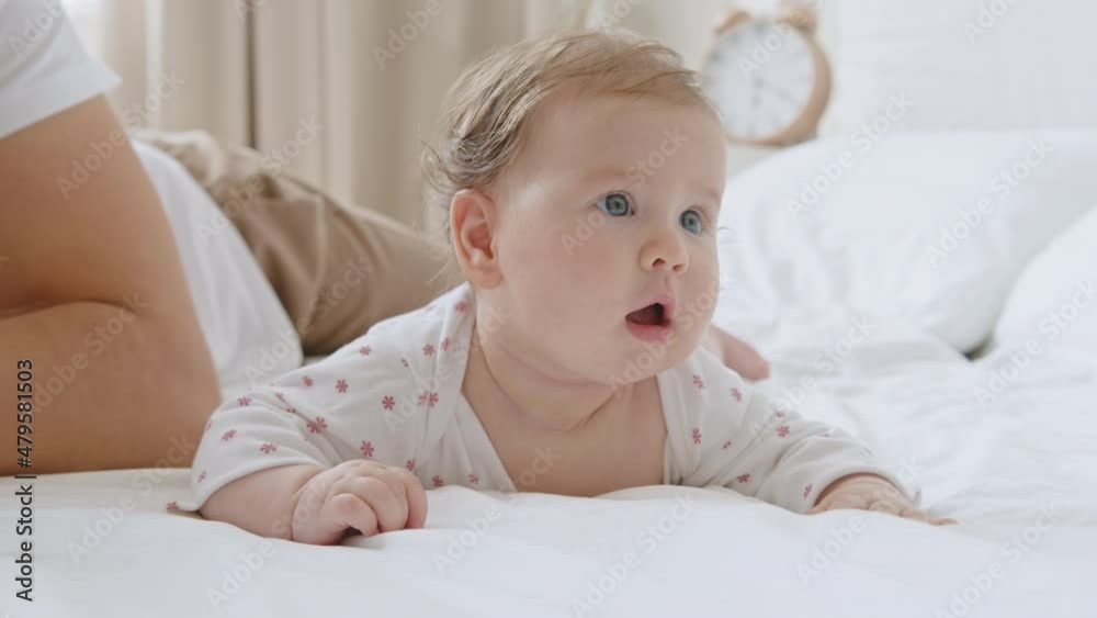 Cute sweet baby newborn with blue eyes lying on white bed sheet on her tummy expressive looks, moving her arms. Third month closeup portrait. Father gently strokes and kiss the child's head Cute sweet baby newborn with blue eyes lying on white bed sheet on her tummy expressive looks, moving her arms. Third month closeup portrait. Father gently strokes and kiss the child's head