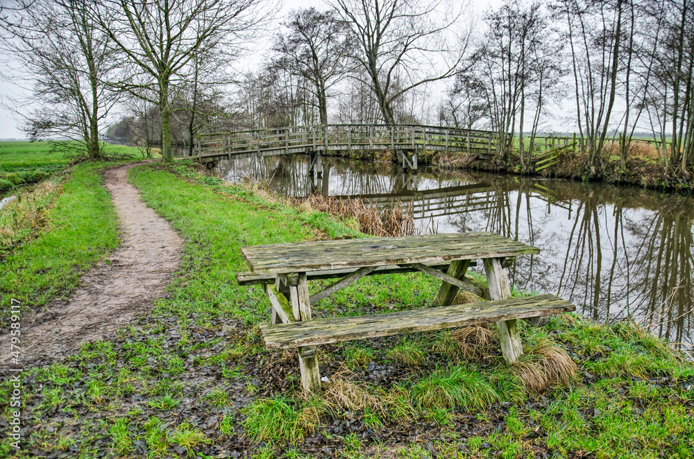 Wooden bench, bridge across a canal and muddy hiking trail in Krimpenerwaard polder in the Netherlands Wooden bench, bridge across a canal and muddy hiking trail in Krimpenerwaard polder in the Netherlands