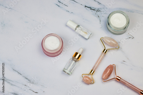 Natural skin care facial massage and relaxation. Anti-aging and lifting concept at home. Face creams, face serum oil, pink quartz roller for face massage on white marble background.