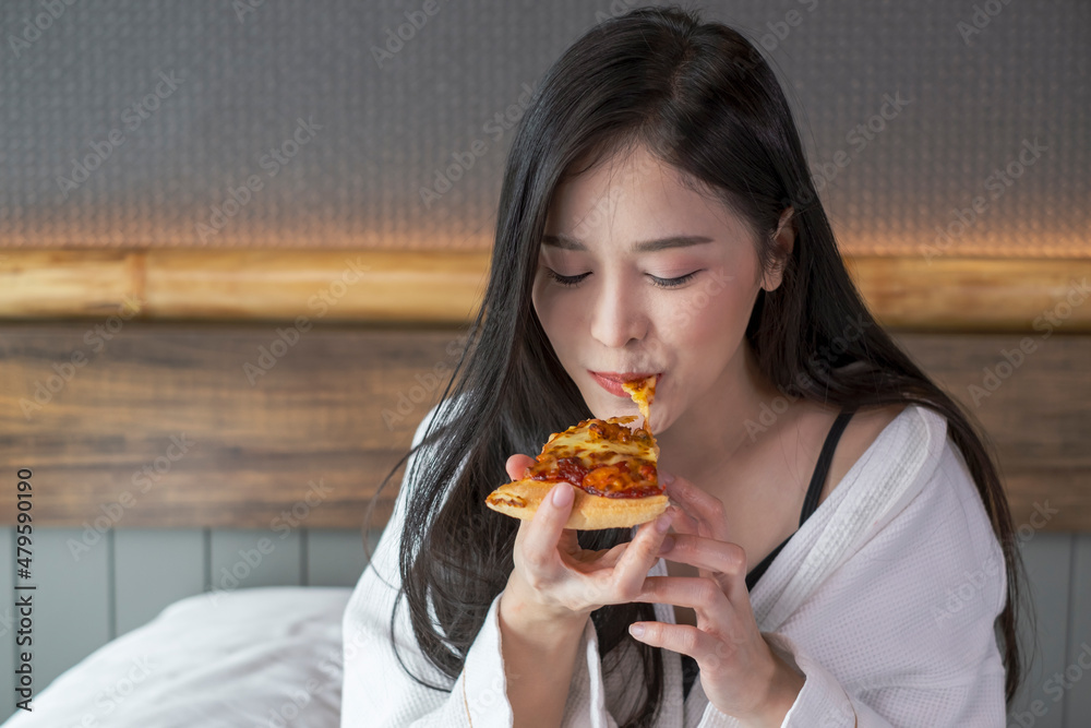 Happy young woman enjoy eating pizza on bed in hotel room. Beautiful female sitting in hotel bed eating pizza. Young woman hanging out eating pizza.