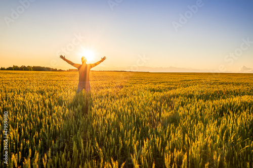 man looking at beautiful sunset and feeling joy in a wheaten shiny field with golden wheat and sun glow on the background, amazing sky and rows of wheat leading far away, valley landscape © Yaroslav