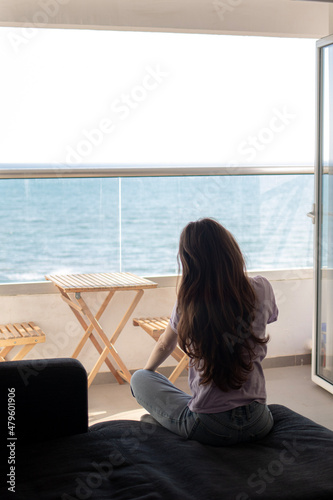 A young woman is sitting on sofa near balcony and looking at the sea