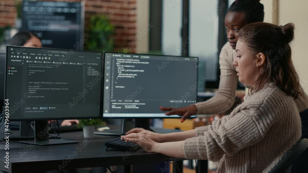 Junior programer writing algorithm under experienced colleague supervision sitting at desk with multiple screens showing programming interface. Developer writing code with the help of colleague.