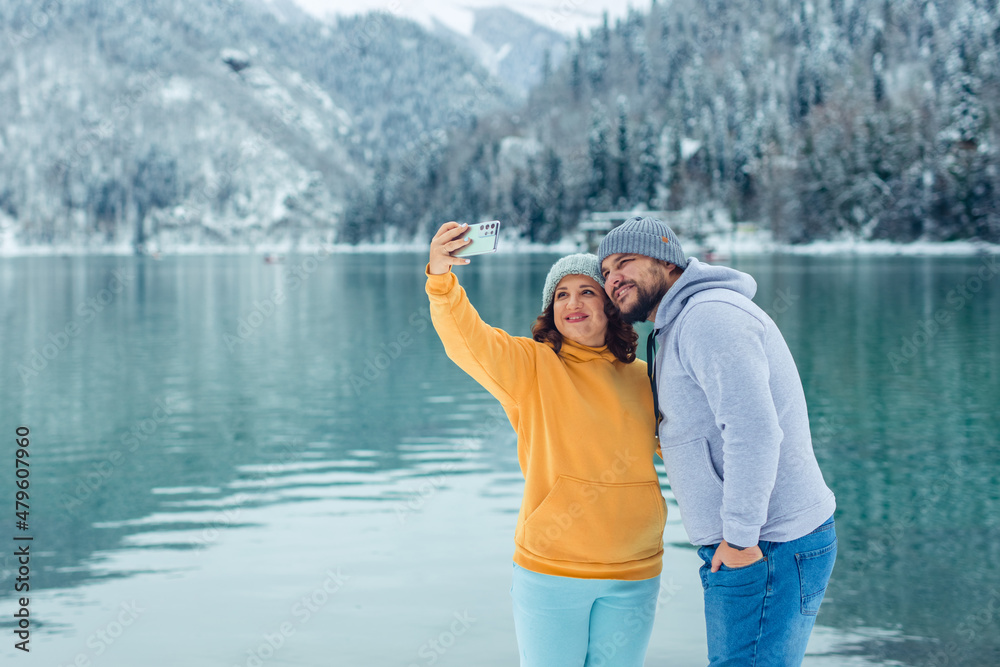 winter travel across Europe. view of the alpine lake with snow. portrait of an adult couple of lovers travelers on the shore of a high-mountain lake taking a selfie on a smartphone