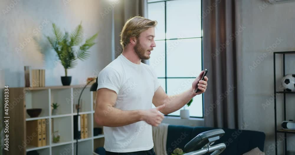 Likable active sportive young bearded man in white t-shirt jogging on treadmill at home and talking with friend via phone video chat Likable active sportive young bearded man in white t-shirt jogging on treadmill at home and talking with friend via phone video chat