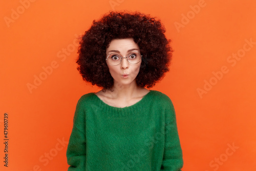 Portrait of funny woman with Afro hairstyle wearing green casual style sweater and eyeglasses, standing with pout lips, looking at camera. Indoor studio shot isolated on orange background.