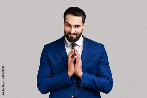 Cunning bearded man clasping hands and planning evil tricky prank or scheming, cheating with sly smile, wearing official style suit. Indoor studio shot isolated on gray background.